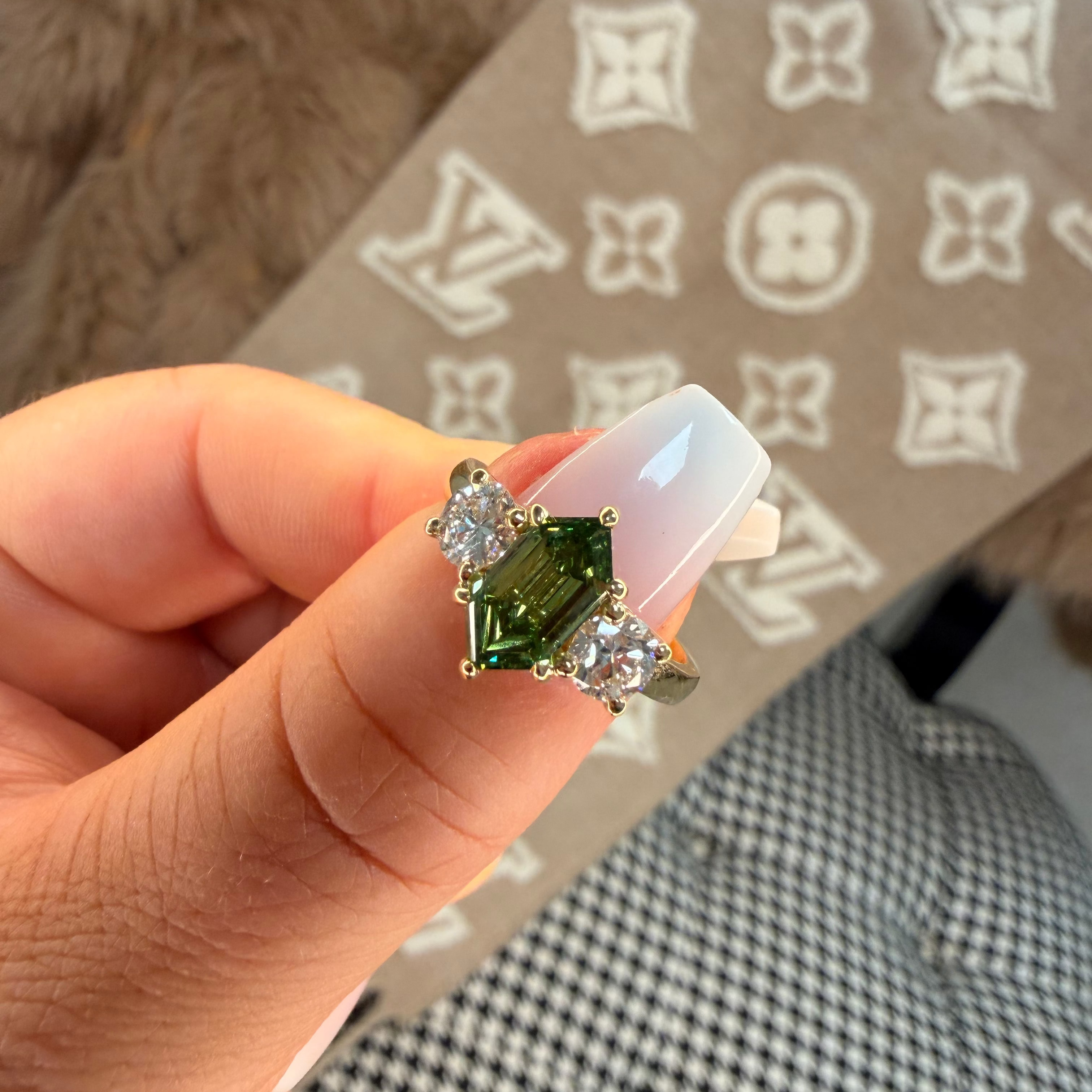 Hand holding a ring with green gemstone and clear stones against a patterned background