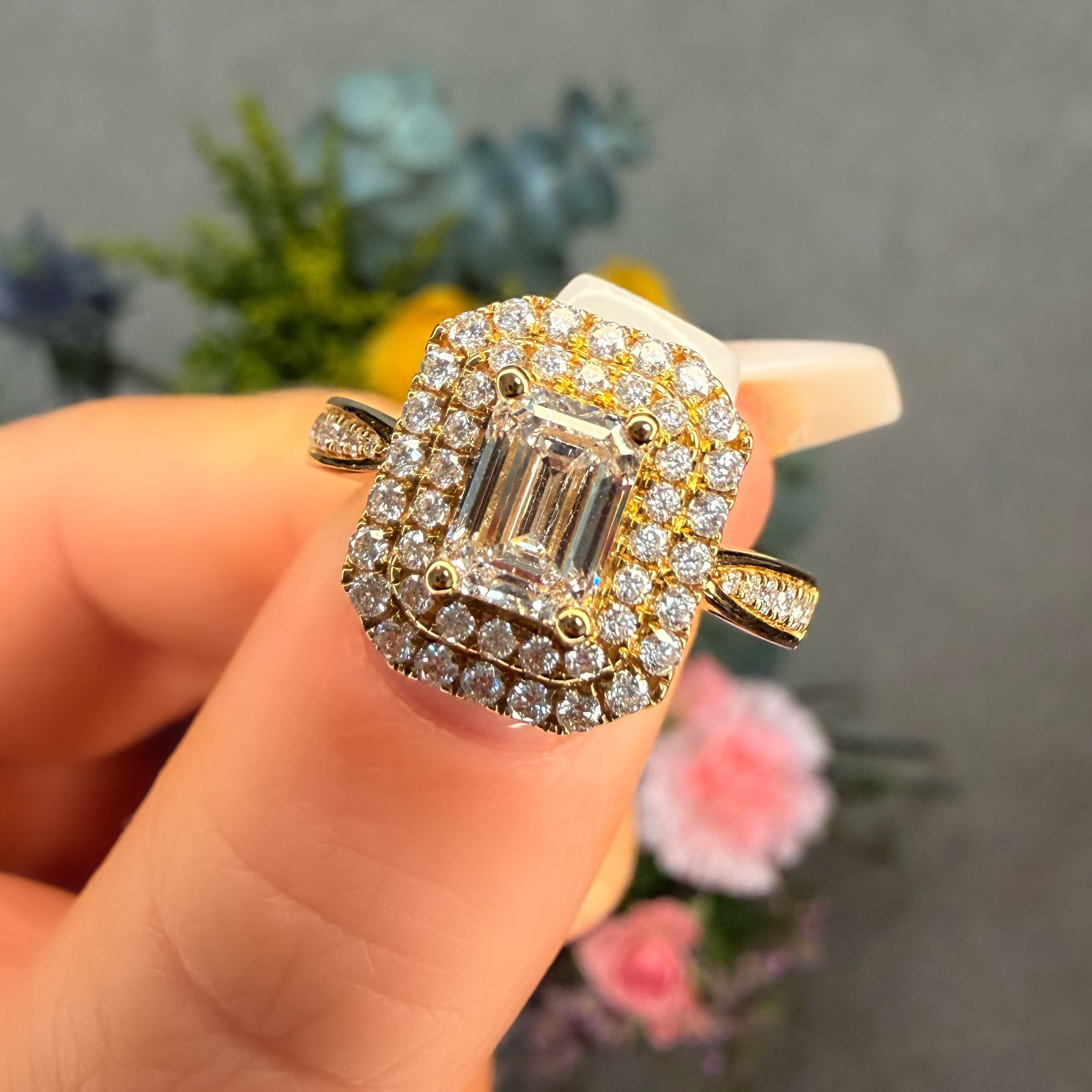 Gold ring with a large diamond held by a hand against a blurred floral background