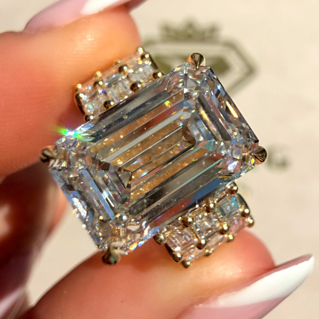 A close-up of a ring being held between fingers. The ring features a large emerald-cut clear center stone with smaller clear accent stones in a gold-tone setting.