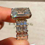 A close-up of a gold ring with a large rectangular clear gemstone and a multi-row baguette setting, held between fingers against a blurred light background.