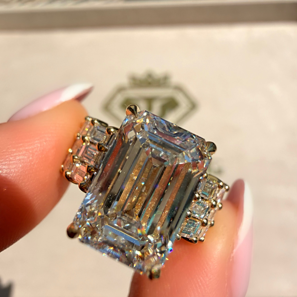 A close-up of a ring being held between fingers. The ring features a large emerald-cut clear center stone with smaller baguette and round side stones in a yellow gold setting.