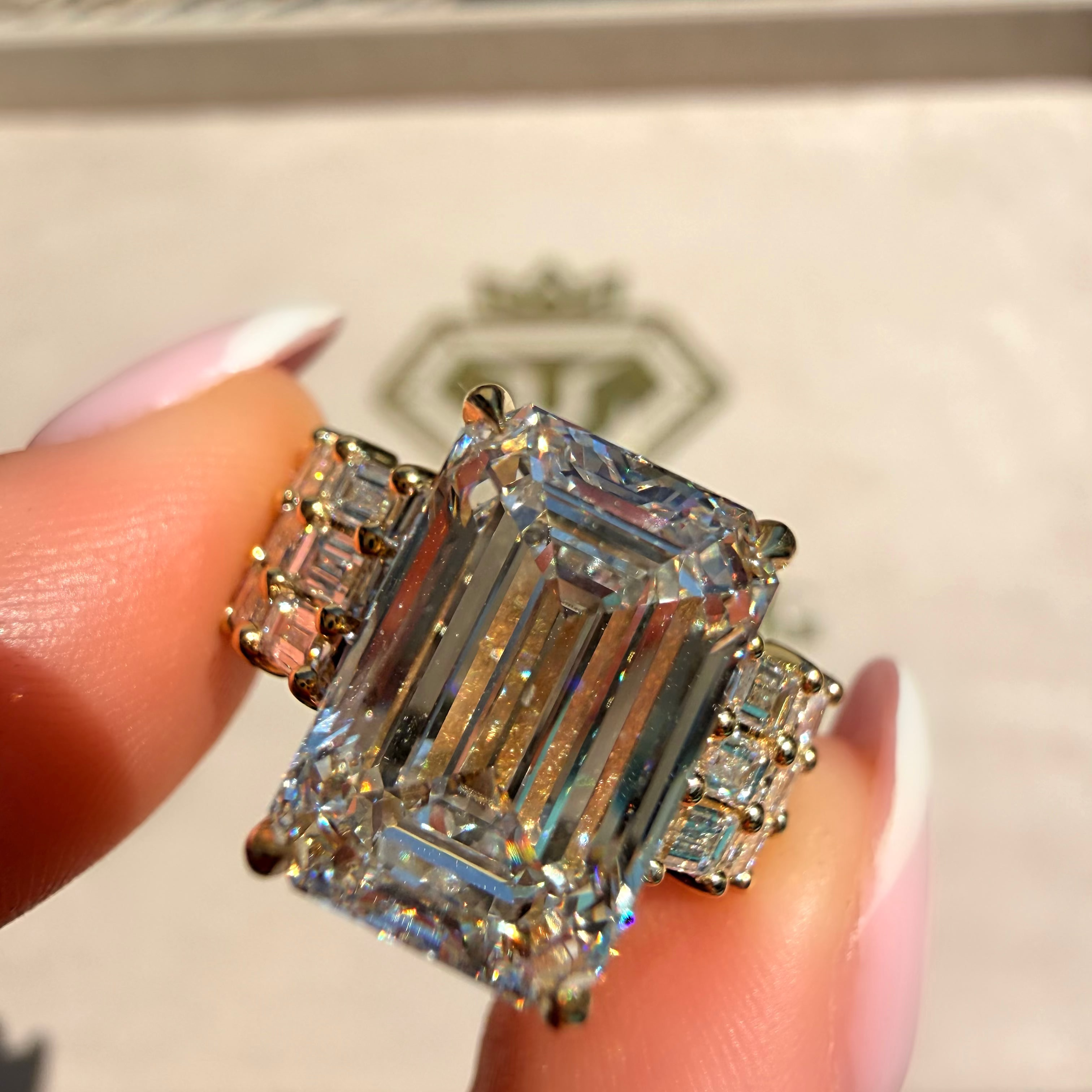 A close-up of a ring being held between fingers. The ring features a large emerald-cut clear center stone with smaller baguette and round side stones in a yellow gold setting.