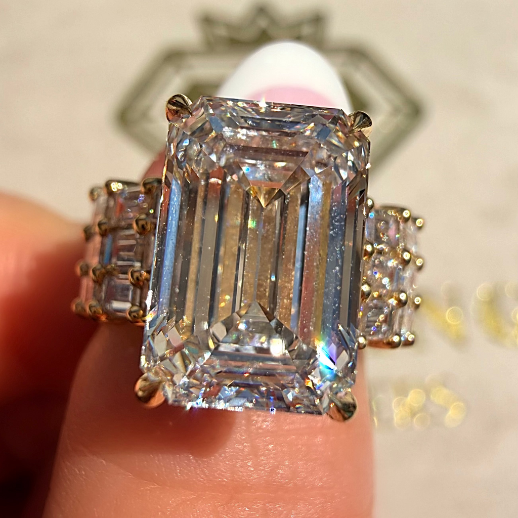 An emerald-cut diamond ring with yellow gold side stones, held between fingers to show scale.