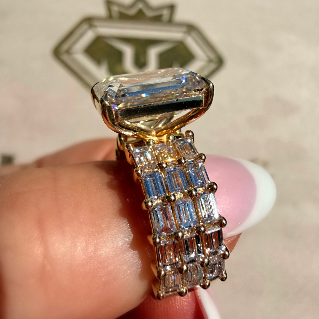 A gold ring with a large rectangular clear gemstone and a multi-row band of baguette-cut clear stones, held between fingers against a light beige background with a blurred logo.