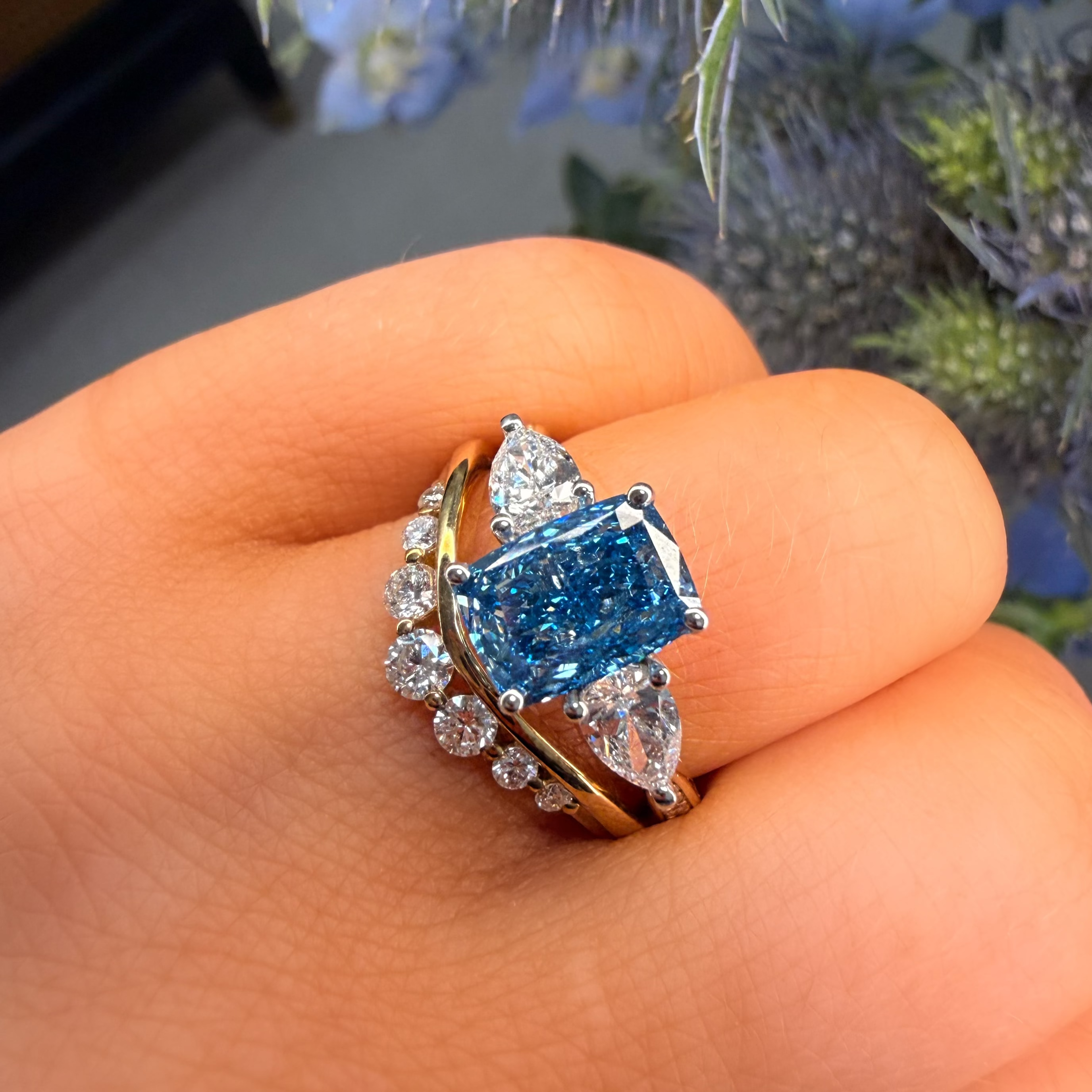 Ring with a blue gemstone and diamond accents on a hand, with a blurred natural background.