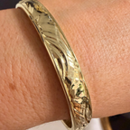A close-up of a gold bangle bracelet with a textured, wavy pattern, worn on a person's wrist.