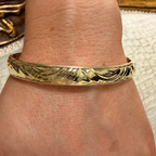 A close-up of a gold bangle bracelet with an engraved leaf pattern, worn on a person's wrist.
