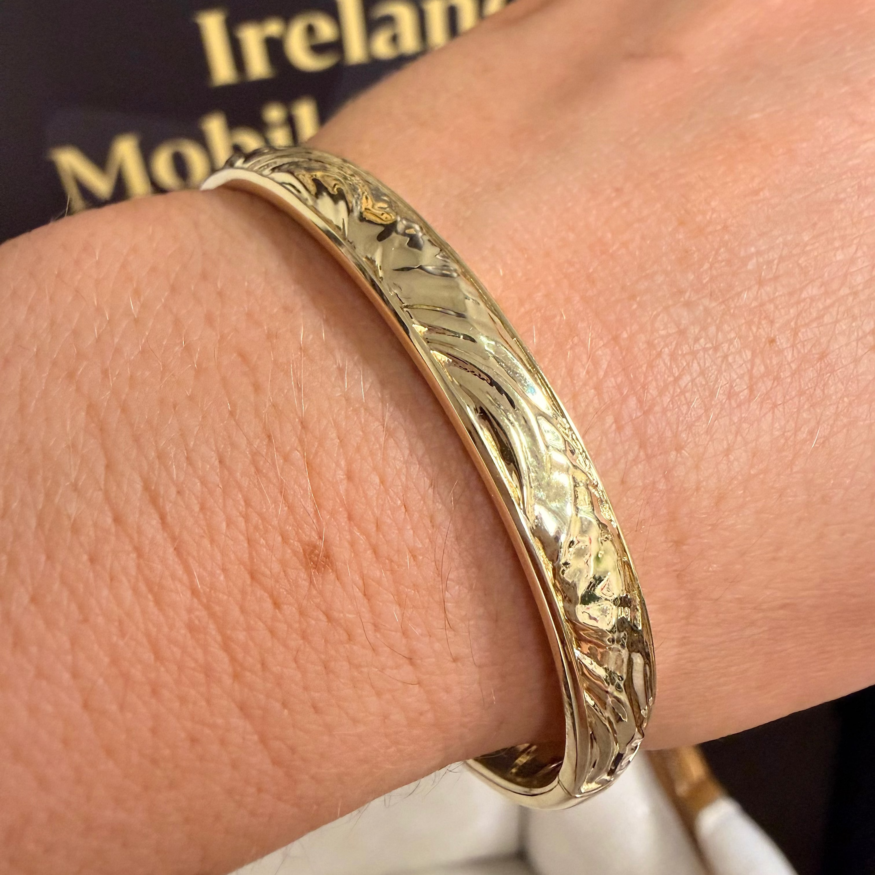 A close-up of a gold bangle bracelet with an engraved floral pattern, worn on a person's wrist. A gold ring is also visible on the finger, and a dark background with gold text is in the upper left.