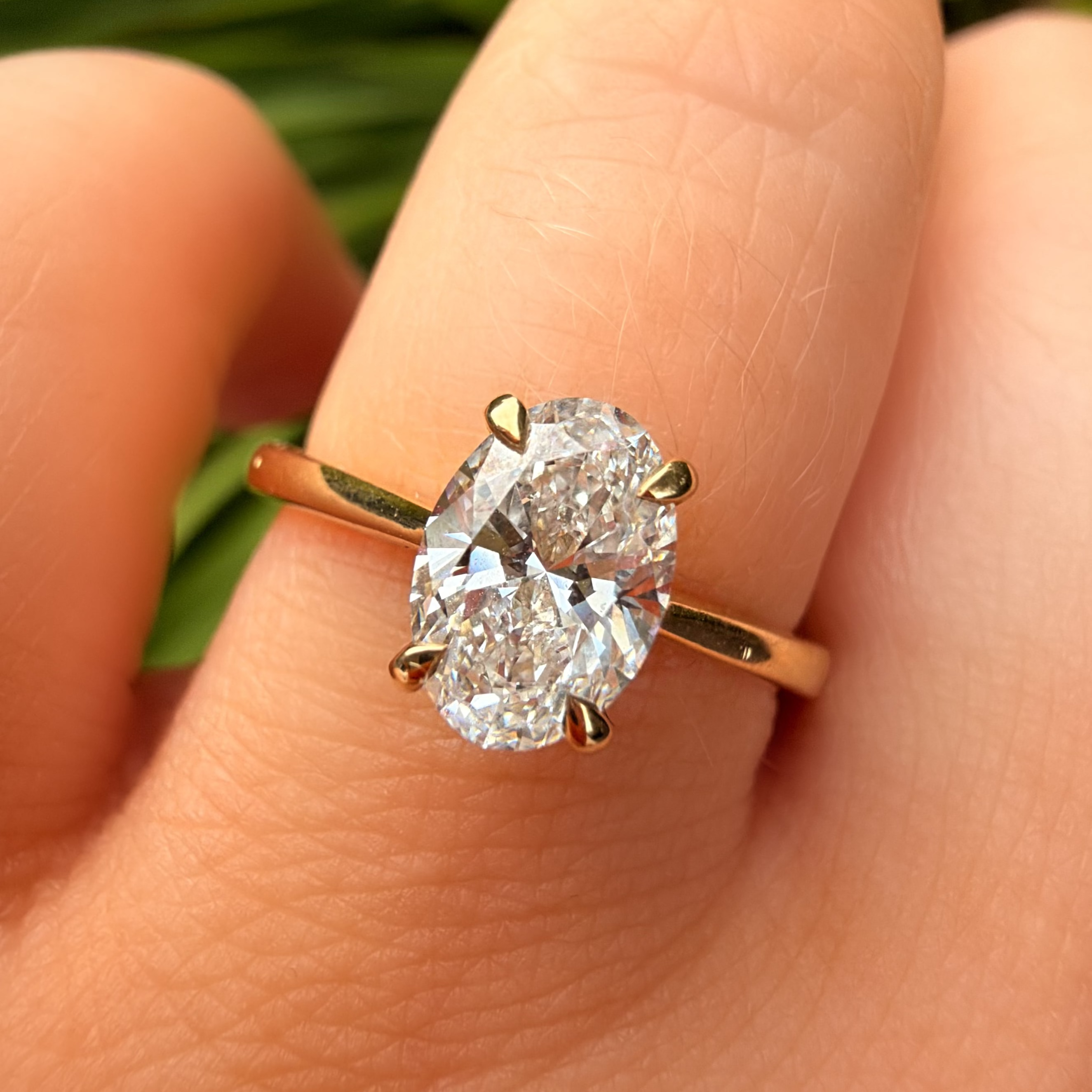A close-up of a hand wearing a yellow gold ring with a large oval-cut clear gemstone in a prong setting, with blurred green foliage in the background.