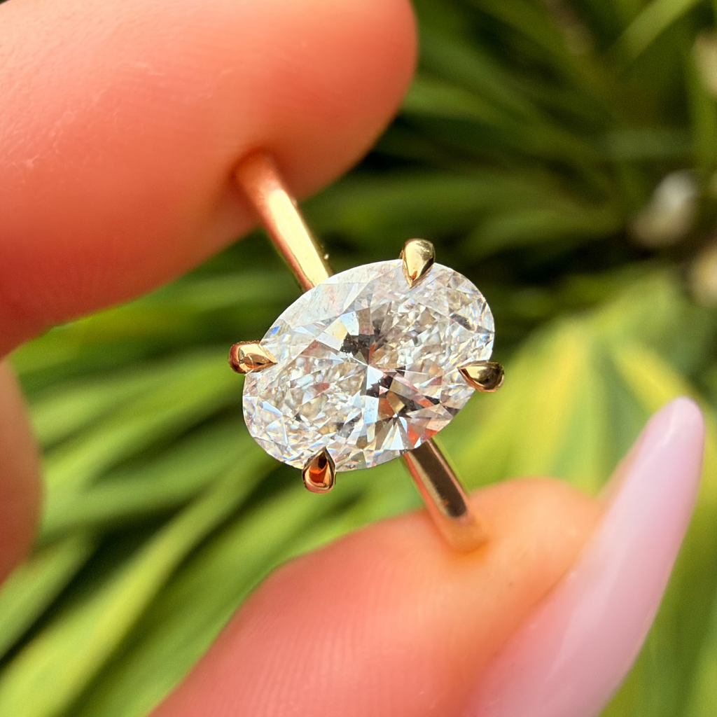A close-up of a hand holding a gold ring with an oval-cut clear gemstone, set against a blurred green outdoor background.
