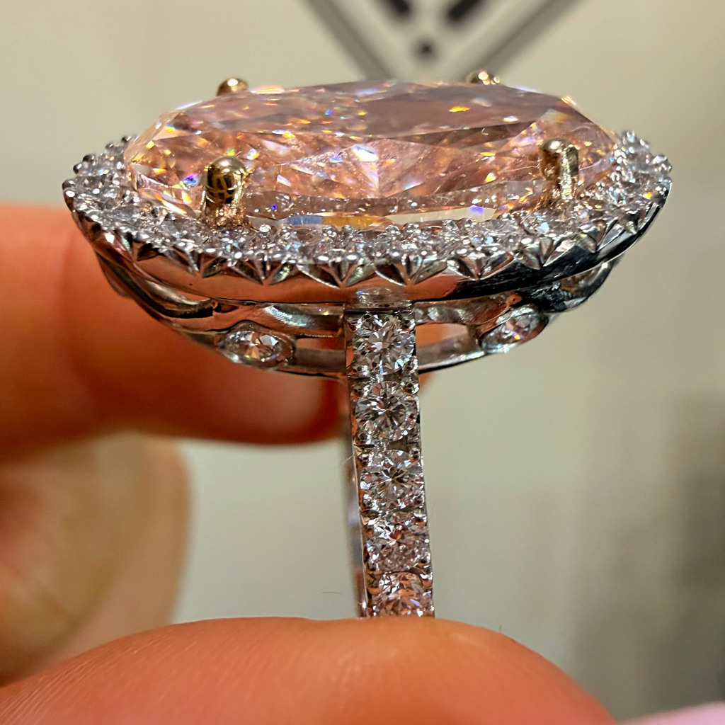 A close-up of a hand holding a ring with a large oval pink gemstone surrounded by a halo of small clear stones and a diamond-set band.