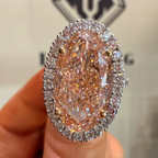 A close-up of a hand holding a large oval pink diamond ring with a halo of small clear stones. The background is a light beige wall with a blurred logo.