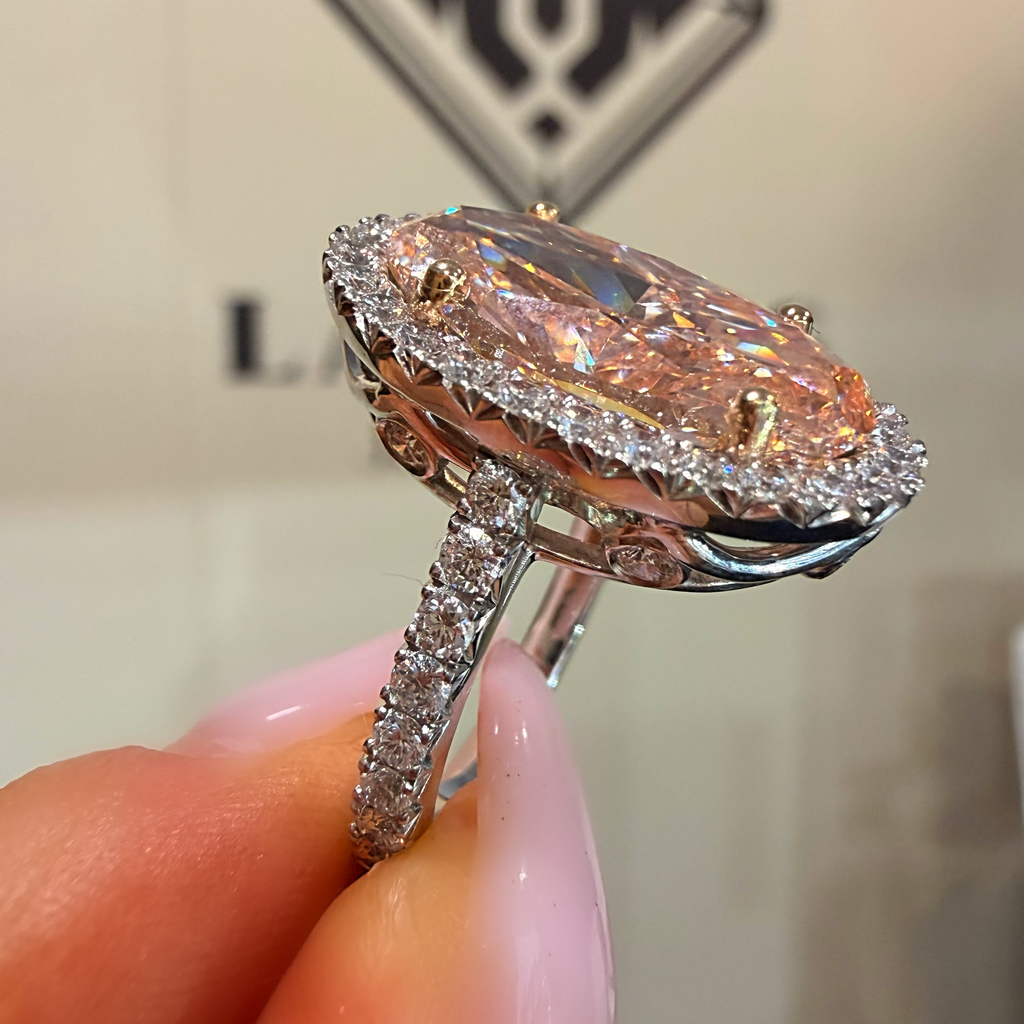 A close-up of a hand holding a silver halo ring with a large marquise-shaped iridescent pink gemstone and small clear accent stones.