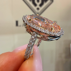 A close-up of a hand holding a silver halo ring with a large marquise-shaped iridescent pink gemstone and small clear accent stones.