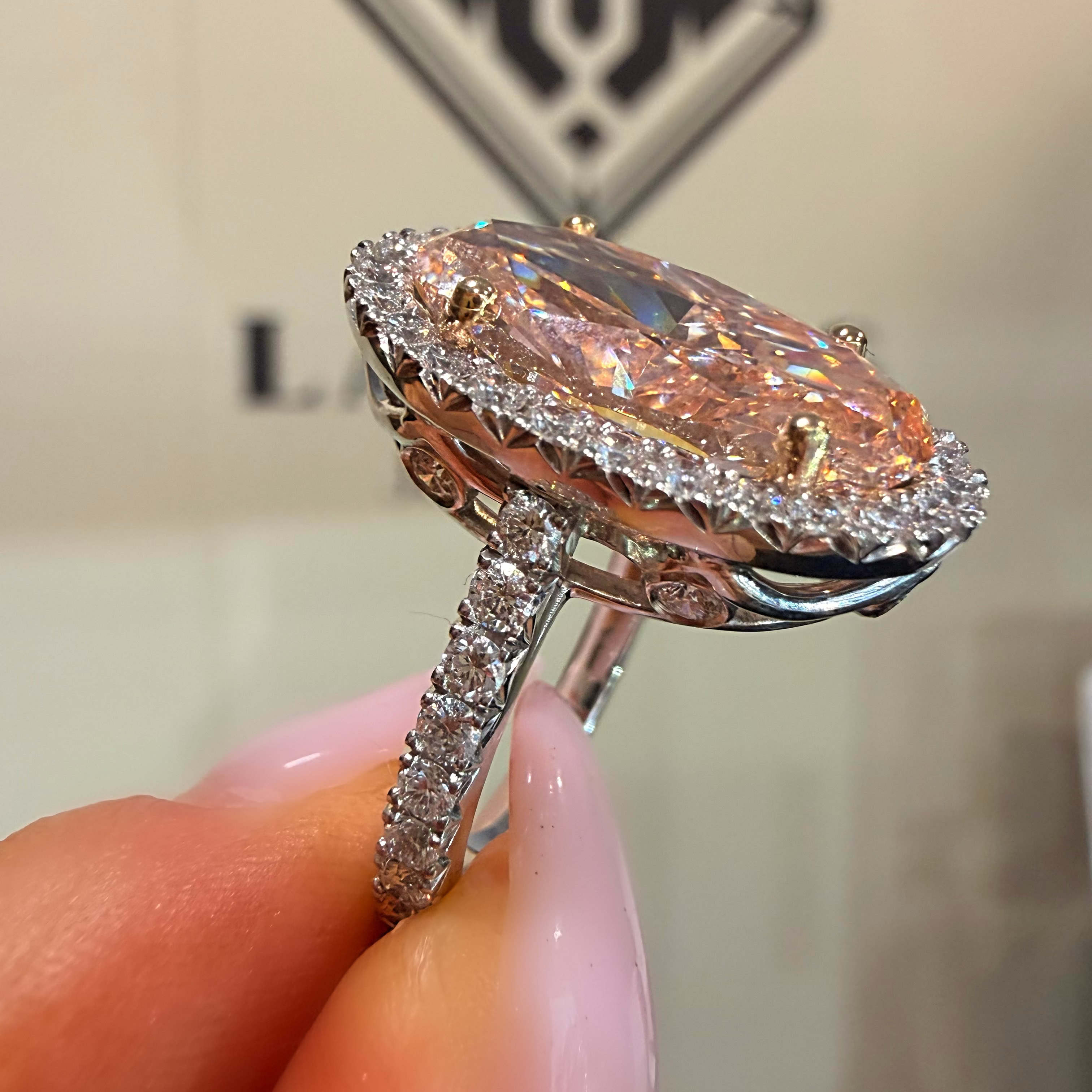 A close-up of a hand holding a silver halo ring with a large marquise-shaped iridescent pink gemstone and small clear accent stones.