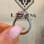 A close-up of a hand with pink manicured nails holding a silver-toned ring featuring a pink gemstone and small clear accent stones, with a blurred jewelry box in the background.