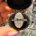 A silver-toned ring with a large oval, highly faceted clear gemstone held in an open black velvet jewelry box with gold trim, being held by a hand with pink manicured nails.