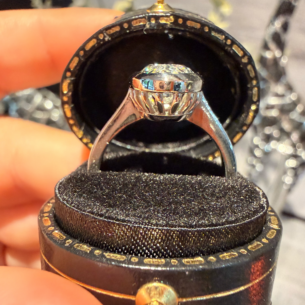 A silver-toned diamond ring with a round brilliant-cut center stone is displayed inside an open black velvet ring box with gold trim, held by a hand.