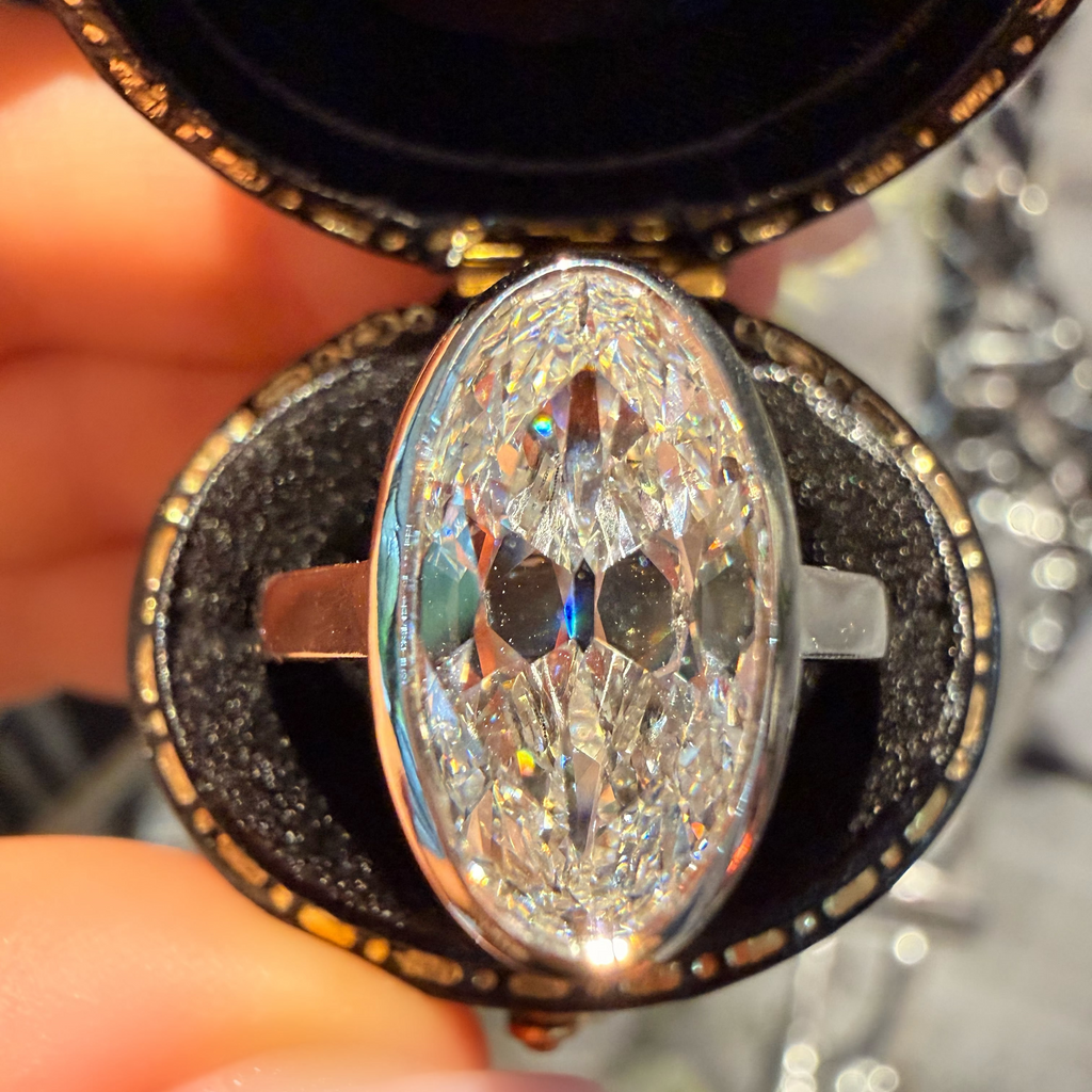 A close-up of a hand holding an open round jewelry box containing a large oval clear gemstone ring with a silver-toned band.