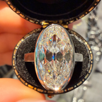 A close-up of a hand holding an open round jewelry box containing a large oval clear gemstone ring with a silver-toned band.
