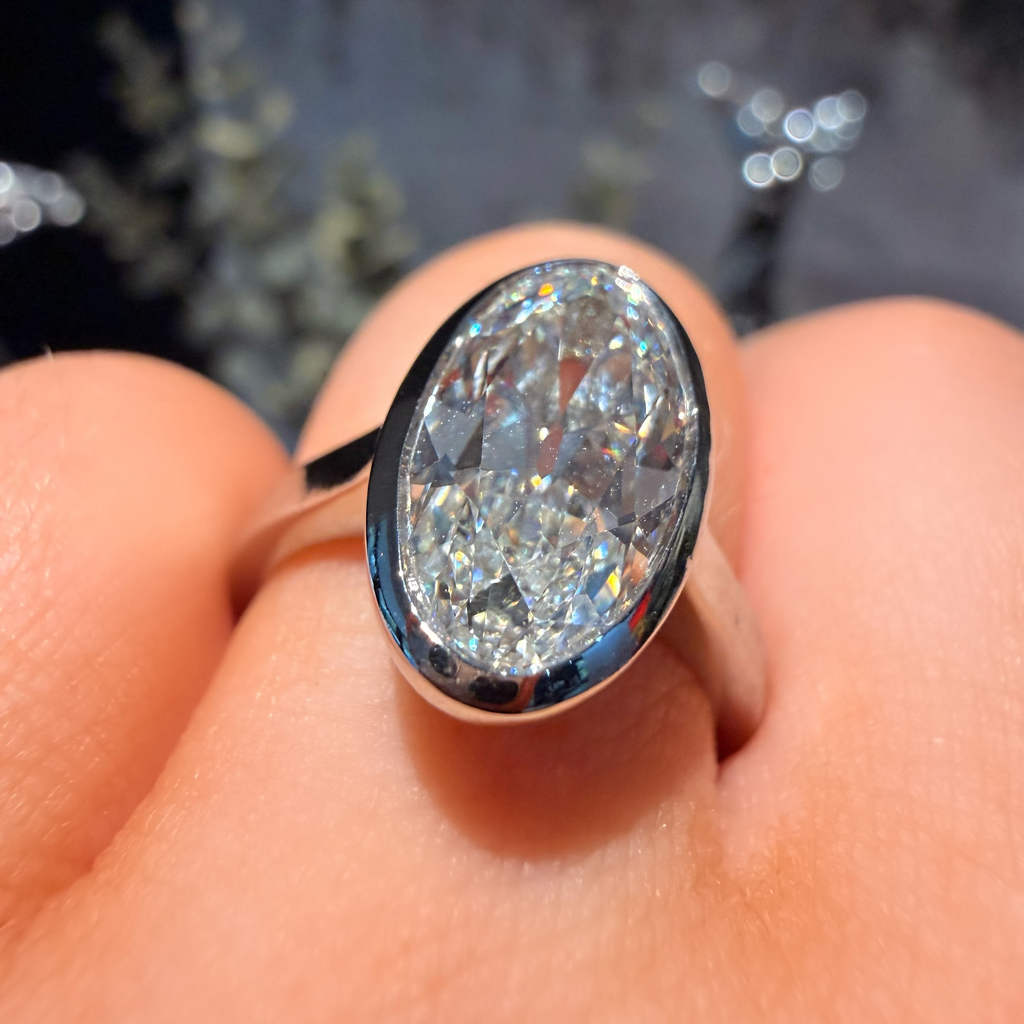 A silver ring featuring a large, oval-cut clear gemstone, worn on a finger. The ring is photographed in close-up against a dark, blurred background with bokeh lights.