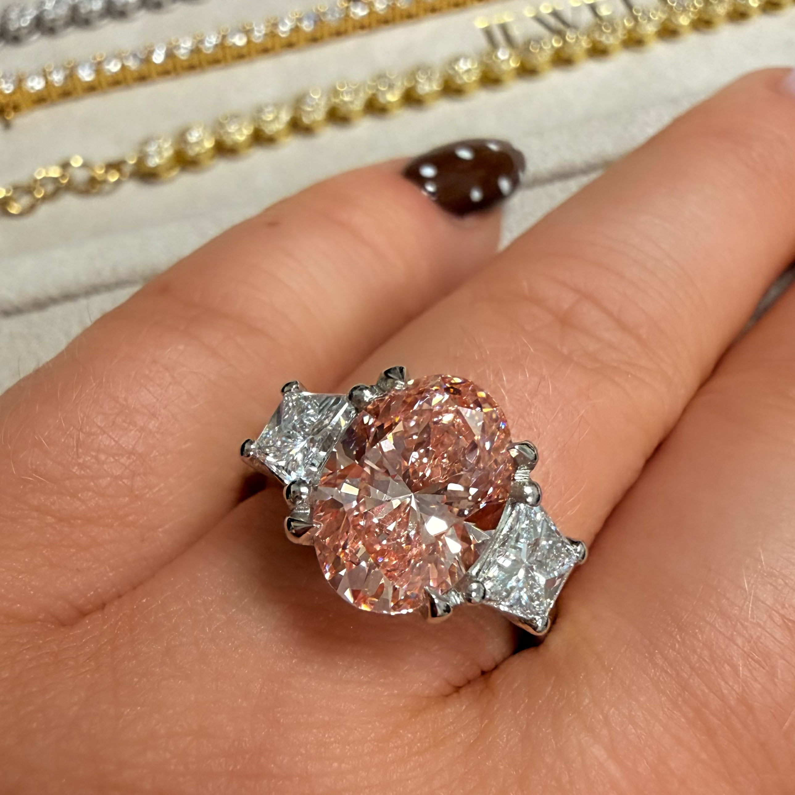 A close-up of a hand wearing a ring with three round pinkish diamonds set in silver-toned metal, with other gold and silver rings blurred in the background.