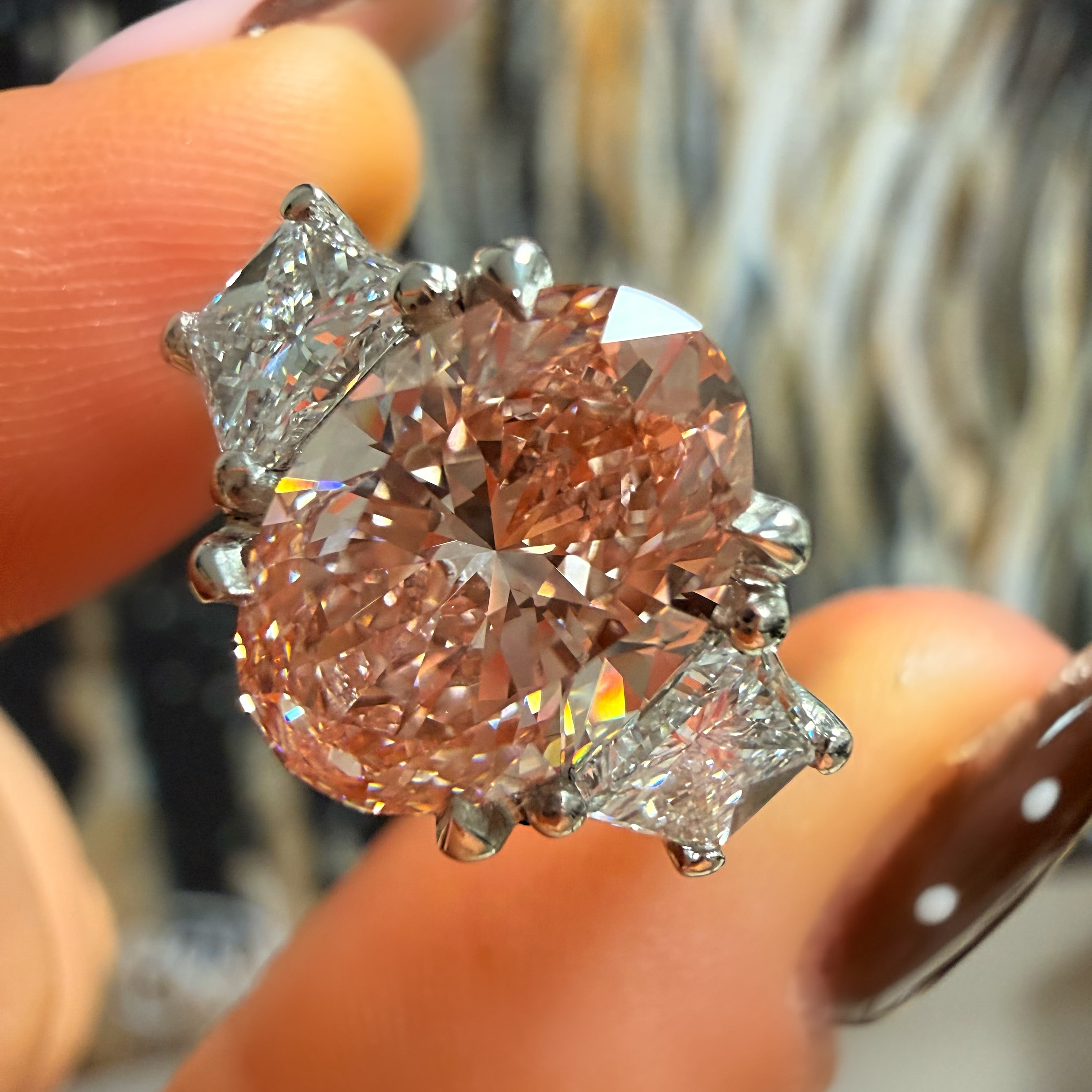 A close-up of a ring held between fingers. The ring features a large round rose-gold center stone with three smaller clear accent stones in a silver-toned setting.