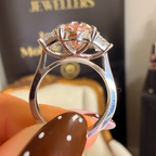 A close-up of a silver-toned ring with a central peach-pink gemstone held by a hand with brown nail polish and white polka dots, with a blurred black LABKING JEWELLERS box in the background.