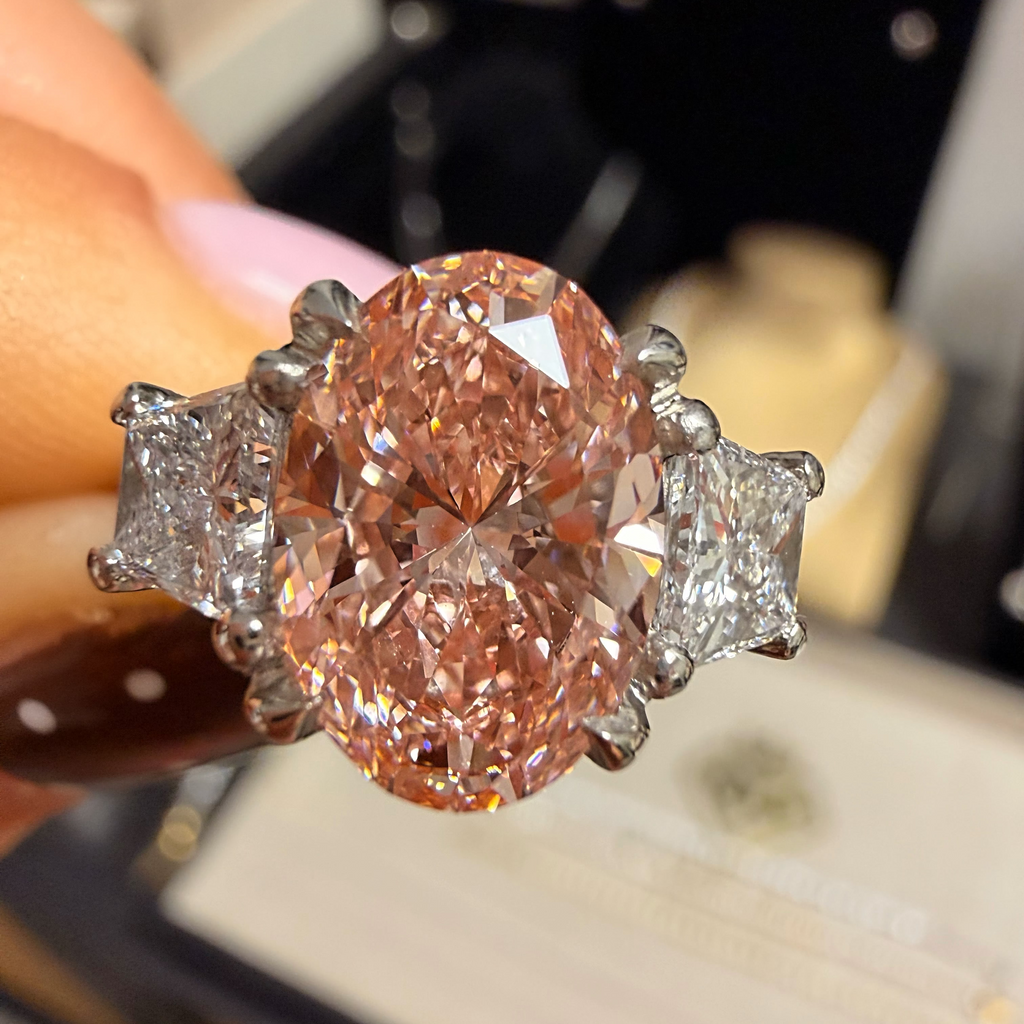 A close-up of a ring held by a hand, featuring a large oval pinkish-orange center stone with two small clear side stones in a silver-toned setting.