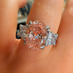 A close-up of a hand wearing a ring with a large oval pinkish gemstone and two smaller clear stones on either side, set in a silver-toned metal.