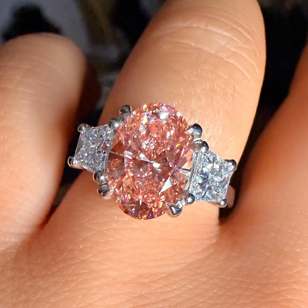 A close-up of a ring worn on a finger, featuring a large oval pink center stone with two small clear side stones in a silver-toned setting.