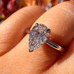 A pear-cut diamond ring set in a silver-colored band, worn on a finger. The diamond is held in four prongs and features a brilliant cut with sparkling facets.