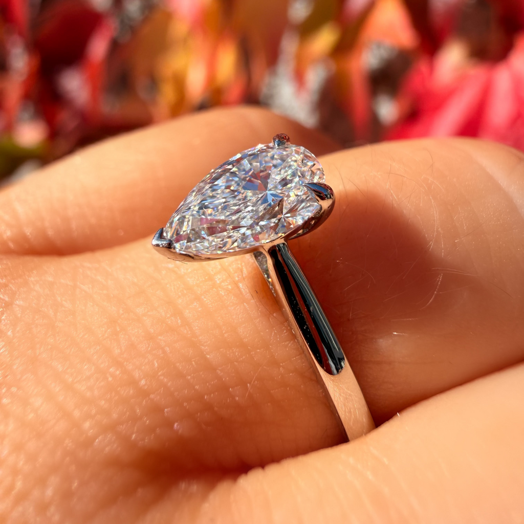 A pear-cut diamond ring in a silver-toned setting, shown worn on a person's finger with a blurred background of red and orange autumn leaves.