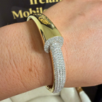 A close-up of a gold hinged bangle bracelet with a pavé-set silver-toned curved accent, worn on a person's wrist. The background shows a dark sign with gold lettering and a light-colored chair.