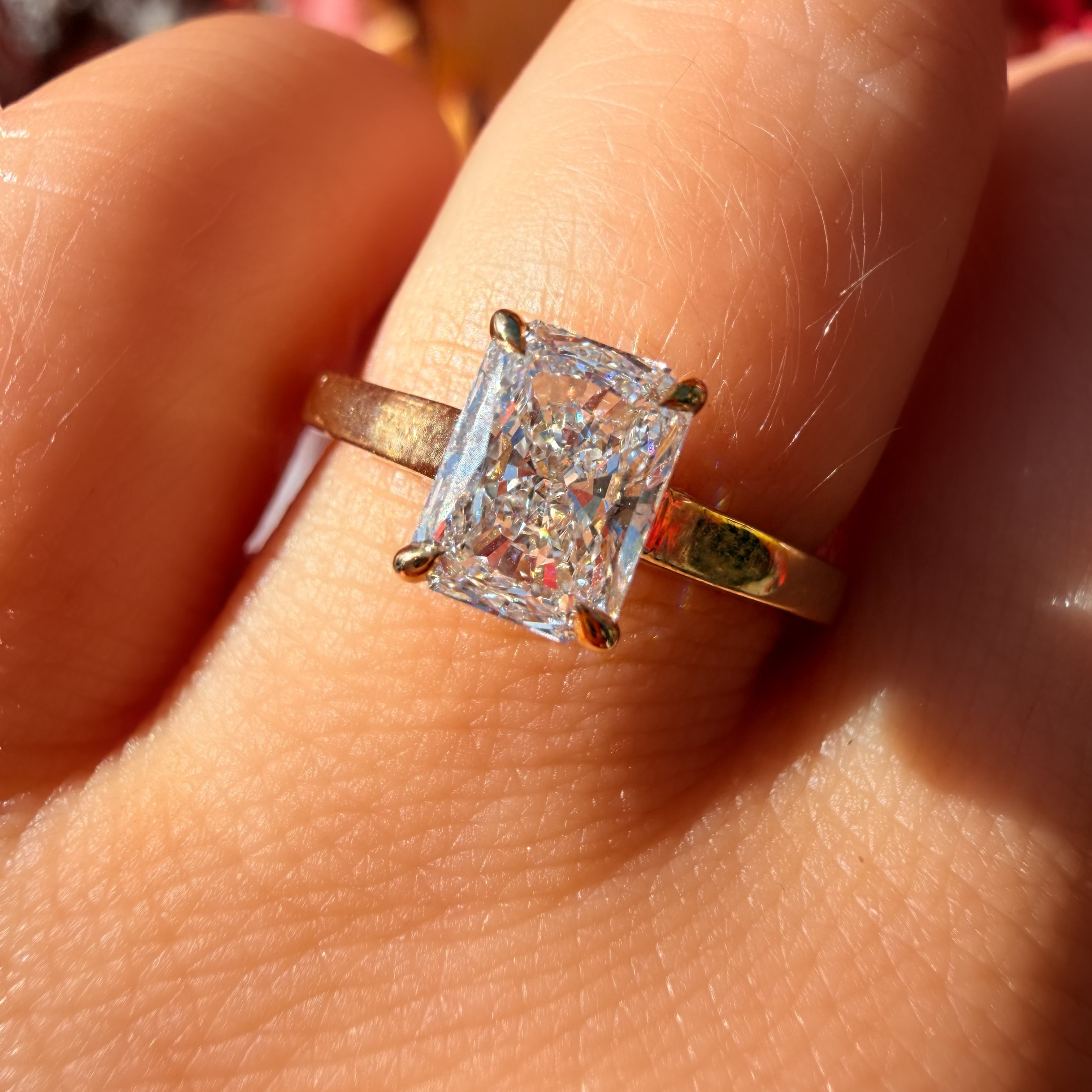 A close-up of a hand wearing a yellow gold ring with a rectangular clear gemstone, set against a blurred background of autumn leaves.