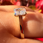 A close-up of a gold ring with a rectangular clear gemstone worn on a finger, set against a blurred background of red and orange autumn leaves.