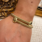 A close-up of a gold-toned double-strand bracelet worn on a person's wrist. The bracelet features one smooth round ball end and one textured snake-like end, set against a background with a decorative gold object and a white textured surface.