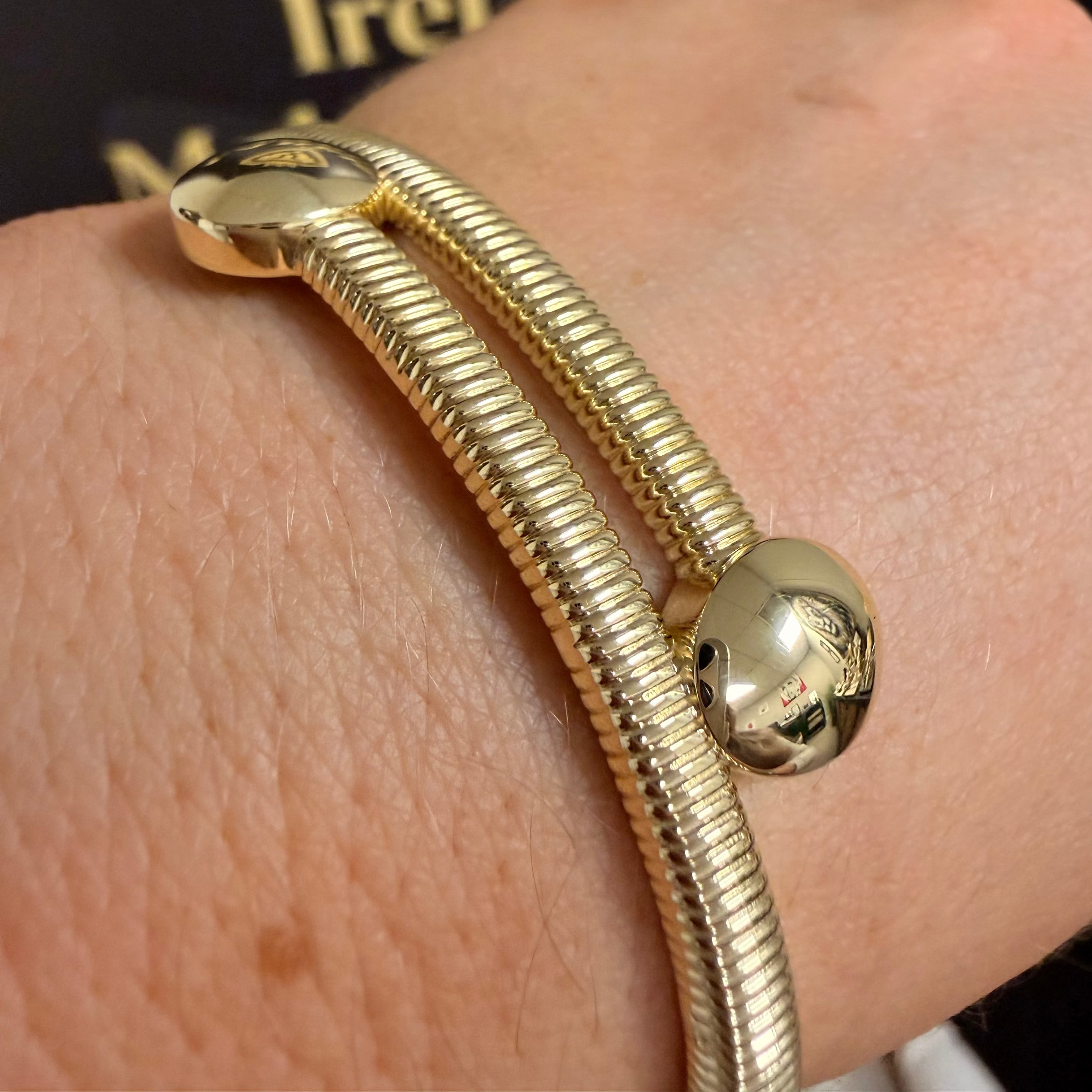 A close-up of a gold-toned double-strand bangle bracelet worn on a person's wrist, with a dark jewelry box in the background.
