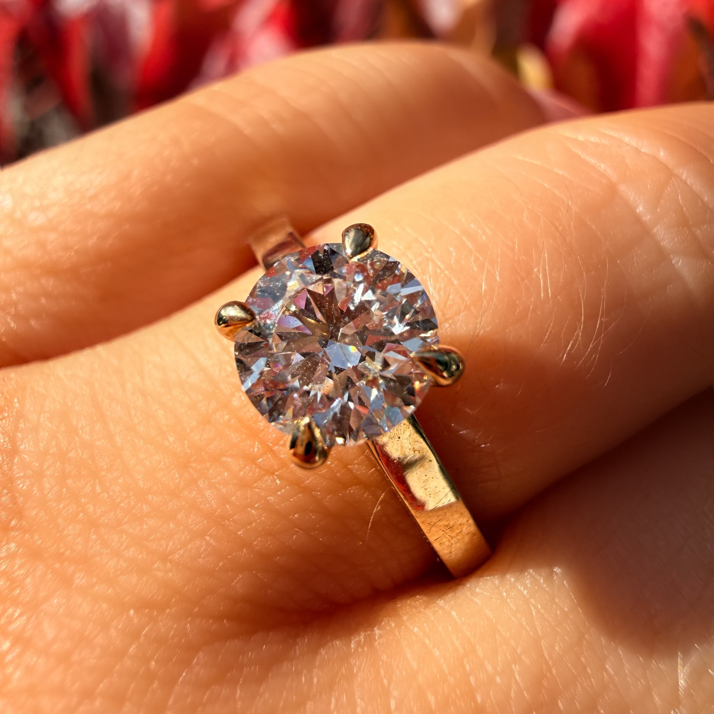 A gold solitaire engagement ring with a round brilliant-cut diamond set on a person's finger, photographed outdoors with a blurred background of red and orange foliage.