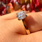 A close-up of a gold ring with a large round brilliant-cut clear gemstone worn on a finger, set against a blurred background of red and orange autumn leaves.