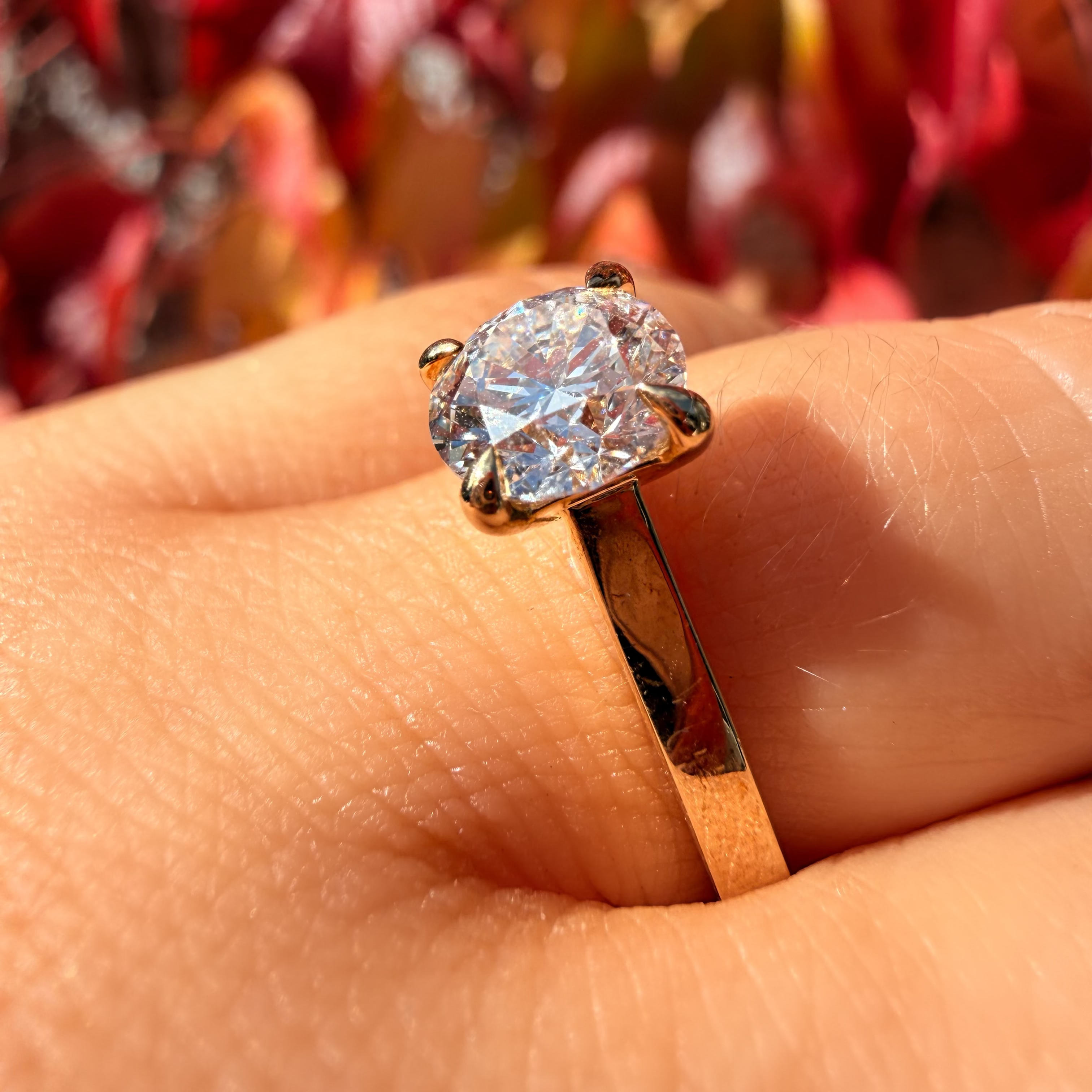 A close-up of a gold ring with a large round brilliant-cut clear gemstone worn on a finger, set against a blurred background of red and orange autumn leaves.
