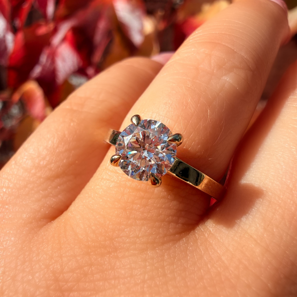 A close-up of a hand wearing a gold ring with a large round clear gemstone, set against a blurred background of red and brown autumn leaves.