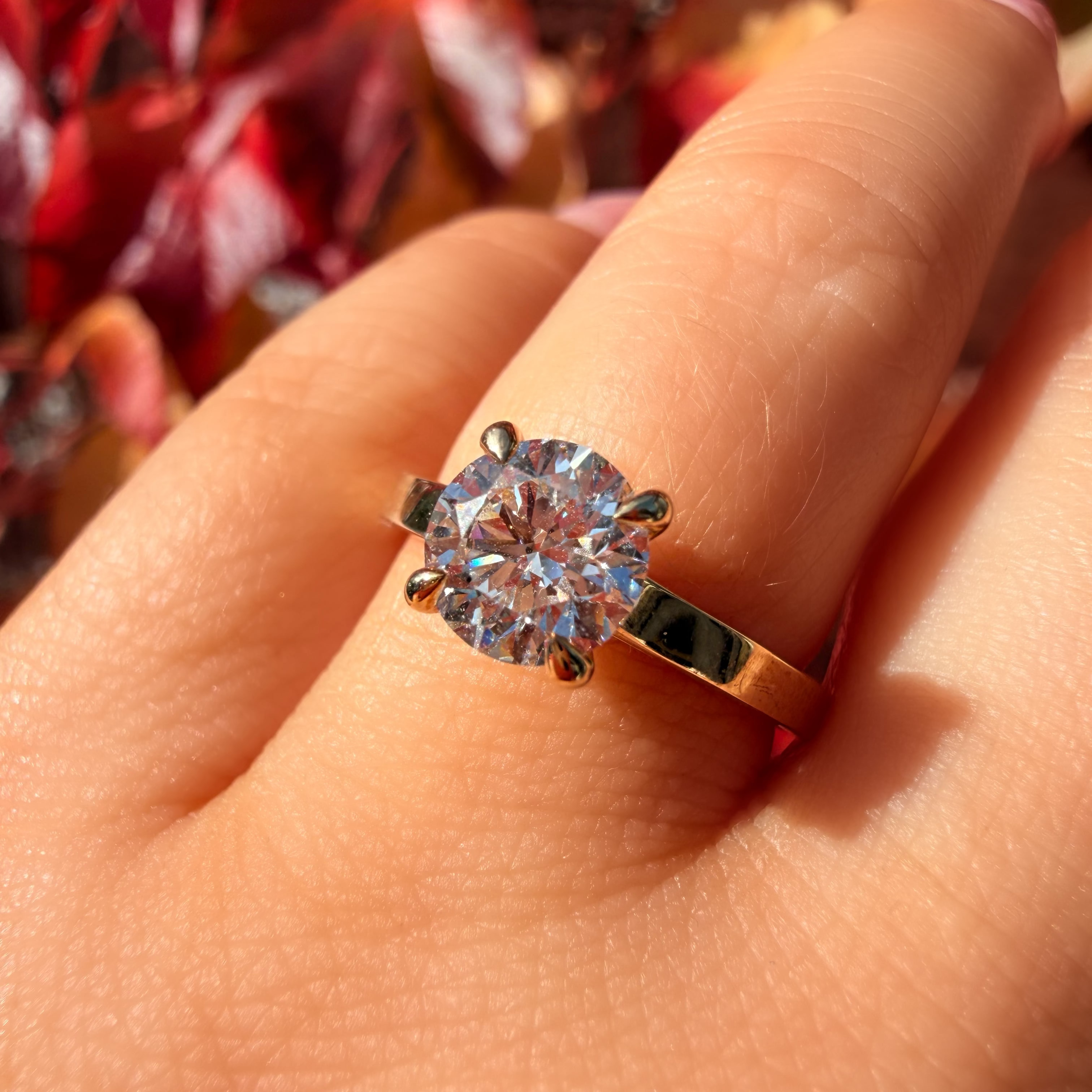 A close-up of a hand wearing a gold ring with a large round clear gemstone, set against a blurred background of red and brown autumn leaves.