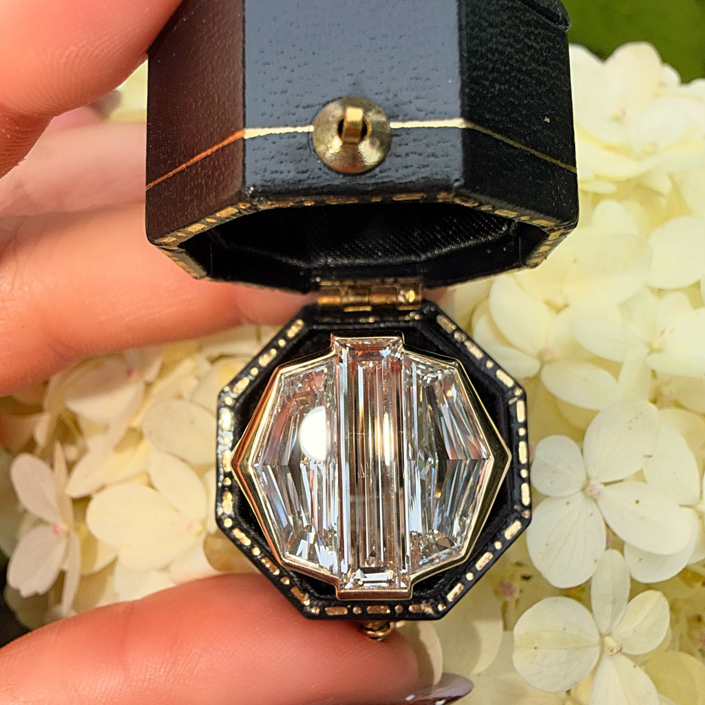 A close-up of a hand holding an open black octagonal jewelry box with gold trim, containing a faceted clear gemstone, set against a background of white flowers.