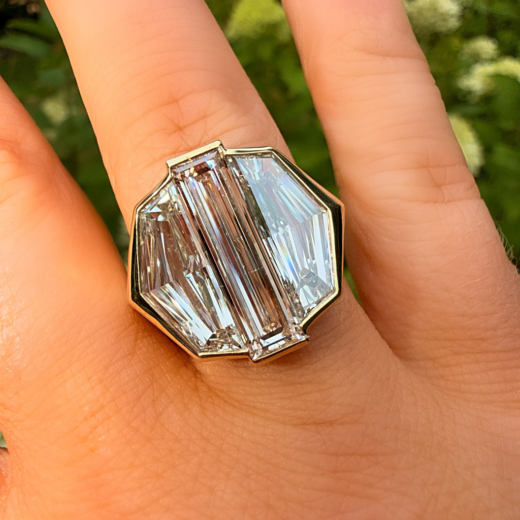 A close-up of a hand wearing a large geometric clear crystal ring with a gold-toned setting, photographed outdoors against a blurred green background.