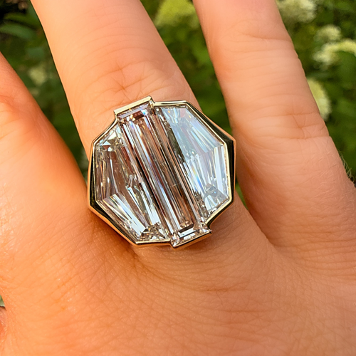 A close-up of a hand wearing a large geometric clear crystal ring with a gold-toned setting, photographed outdoors against a blurred green background.