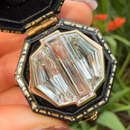 A close-up of a hand holding an open octagonal black and gold jewelry box containing a large, faceted clear baguette-cut gemstone, with a blurred green garden background.