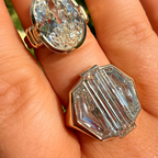 Two Swarovski crystal rings displayed on a hand, featuring an oval-cut crystal in a gold band and an octagonal-cut crystal in a rose gold band, with a blurred green outdoor background.