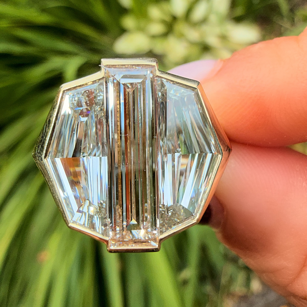 A close-up of a hand holding a gold-toned ring with a large, faceted clear crystal setting against a blurred green garden background.
