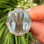 A close-up of a hand holding a gold-toned ring with a large, faceted clear crystal setting against a blurred green garden background.